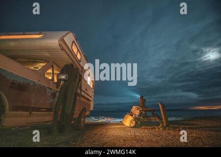 Frau, die auf einem Picknicktisch neben dem Wohnmobil sitzt, während der Surftour Stockfoto