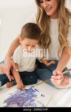 Junge, der mit bunten Farben auf Leinwand mit lächelnder Mutter spielt Stockfoto