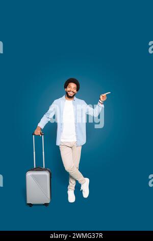 Der temperamentvolle junge Reisende zeigt glücklich zur Seite, steht mit einem Koffer und einem Ausdruck der Freude, schlägt Empfehlungen oder Reiseoptionen vor, isoliert auf Blau Stockfoto