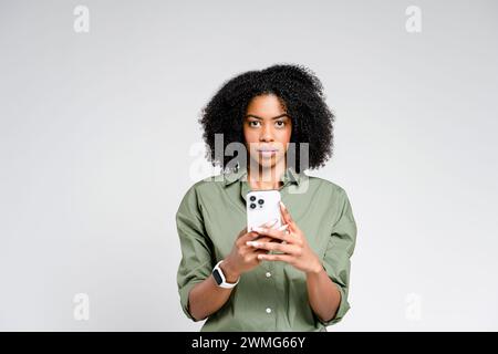 Eine elegante afroamerikanische Frau steht komponiert, hält ein Smartphone in beiden Händen mit einem ernsten Ausdruck und stellt ein professionelles Verhalten dar Stockfoto