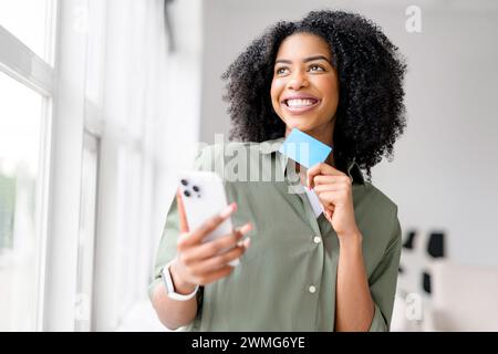 Eine afroamerikanische Frau lacht leicht, während sie auf ihr Smartphone schaut, eine blaue Kreditkarte in der Hand, die auf ein freudiges Online-Shopping-Erlebnis oder die einfache digitale Verwaltung von Finanzen hindeutet. Stockfoto