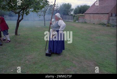 Eine kräftige Frau mit einem Ale-Tankard entspannt ihren Stab-Hecht, um andere Mitglieder der Sealed Knot Society aus dem 17. Jahrhundert zu beaufsichtigen, die im englischen Bürgerkrieg verkleidet sind, feuern eine Kanjon ab, während sie vor Außengästen im The Fleece Inn, Bretforton, Worcestershire, auftreten. an einem Septembertag, an dem die Sealed Knot Society eine Nachstellung des 17. englischen Bürgerkriegs gab Stockfoto