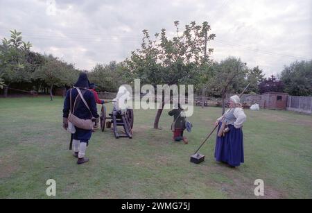 Eine kräftige Frau mit einem Ale-Tankard entspannt ihren Stab-Hecht, um andere Mitglieder der Sealed Knot Society aus dem 17. Jahrhundert zu beaufsichtigen, die im englischen Bürgerkrieg verkleidet sind, feuern eine Kanjon ab, während sie vor Außengästen im The Fleece Inn, Bretforton, Worcestershire, auftreten. an einem Septembertag, an dem die Sealed Knot Society eine Nachstellung des 17. englischen Bürgerkriegs gab Stockfoto