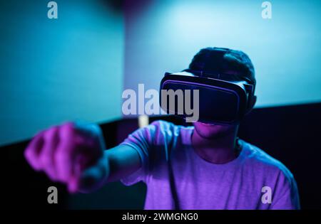 Junger Mann mit einem VR-Headset, der in einem Raum mit violetter Beleuchtung ein immersives Virtual-Reality-Erlebnis macht und verschiedene Handbewegungen in der Luft macht. Stockfoto