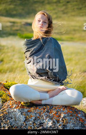 Junge Erwachsene Frau im Yoga-Anzug und gestohlen sitzt in Lotusposition auf einem Stein im grünen Gras und lächelt. Vertikale Zusammensetzung. Stockfoto