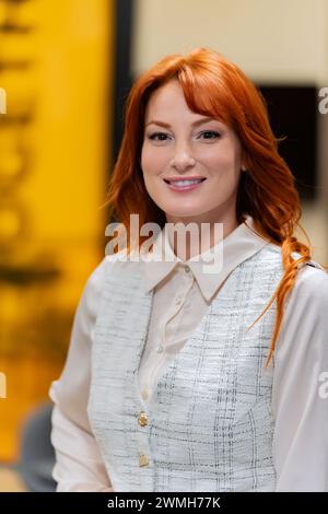 Freundliche rothaarige professionelle Frau, die in einer weißen Bluse und strukturierter Weste lächelt, im Hintergrund des Büros. Stockfoto