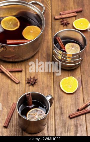 Glühwein mit Zitrusfrüchten und Gewürzen in Metallbecher und Topf. Sternanis, Orangenkeile und Zimtstangen auf dem Tisch. Hölzerner Hintergrund. Stockfoto