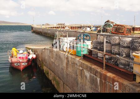 Ein kleines Fischerboot mit einem Mann neben dem Tingwall Pier, mit Angeln und Kisten auf dem Pier und anderen vertäuten Booten, Orkney, Schottland, Großbritannien Stockfoto