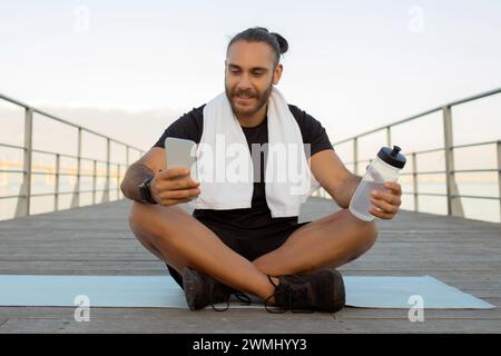 Der sportliche Kerl, der nach dem Training sein Smartphone überprüft, macht eine Pause im Freien Stockfoto