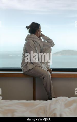 Besinnliche junge Frau, die vom Fenster eines modernen Zimmers aus in bequemer Loungewear auf das Meer blickt Stockfoto