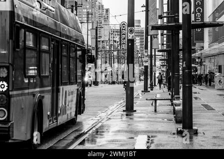 Ein Schwarzweißfoto eines TransLink-Busses, der an einem regnerischen Tag die Granville Street entlang fährt, mit Leuten, die auf dem Bürgersteig mit Sonnenschirmen laufen. Stockfoto