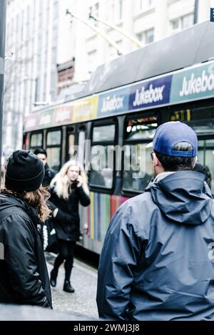 Ein Mann und eine Frau im Vordergrund, die in Richtung eines bunten Trolley-Busses schauen, und eine Frau, die auf ihr Telefon redet und auf sie zugeht. Stockfoto