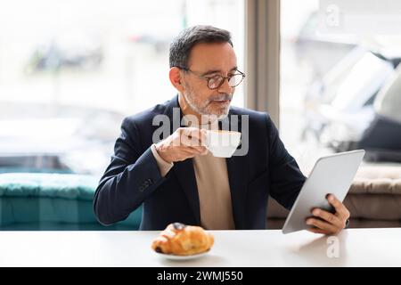 Ein erfahrener reifer Mann in Gläsern schlürft aus einer weißen Espresso-Tasse, während er auf einem Tablet liest Stockfoto