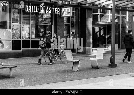 Vancouver, Kanada - 16. Februar 2024: Ein Schwarzweißbild eines Mannes, der mit seinem Fahrrad auf dem Bürgersteig der Granville Street läuft. Stockfoto