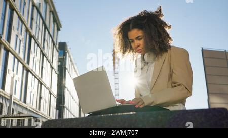 Junge professionelle Frau konzentriert sich auf ihren Laptop-Bildschirm, während sie draußen arbeitet, mit modernen Gebäuden im Hintergrund. Stockfoto