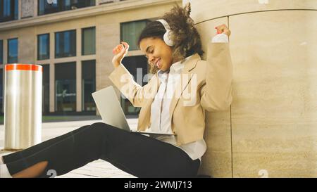Eine junge professionelle Frau genießt eine fröhliche Pause und tanzt zur Musik von ihren Kopfhörern, während sie an ihrem Laptop arbeitet. Sie sitzt im sonnendurchfluteten Ambiente eines städtischen plaza. Stockfoto