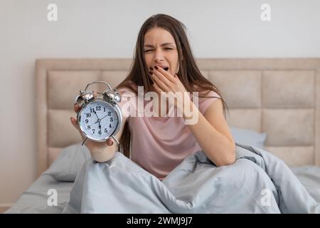 Schläfrige Frau im Bett, die einen Wecker hält und vor Müdigkeit gähnt Stockfoto