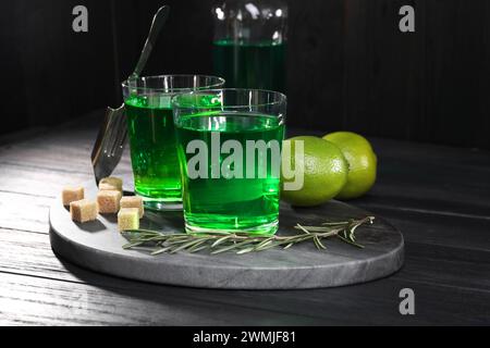 Absinth in Gläsern, Rosmarin, brauner Zucker und Limette auf schwarzem Holztisch. Alkoholisches Getränk Stockfoto