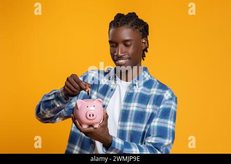 Ein lächelnder junger Schwarzer mit Zöpfen, der Münzen in die Sparkasse einlegt Stockfoto