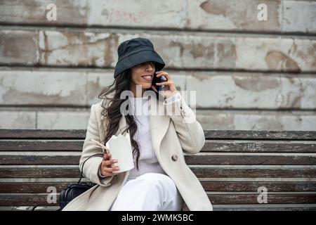 Brünette, die telefoniert, isst chinesische Nudeln und sitzt mit Hut auf der Straße in der Stadt Stockfoto