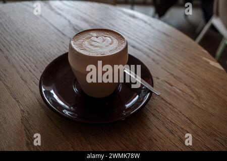 Ein frisch gebrühter Latte mit einer herzförmigen Kunst auf dem Schaum, der auf einer dunkelbraunen Keramikuntertasse und einer Holztheke sitzt. Stockfoto