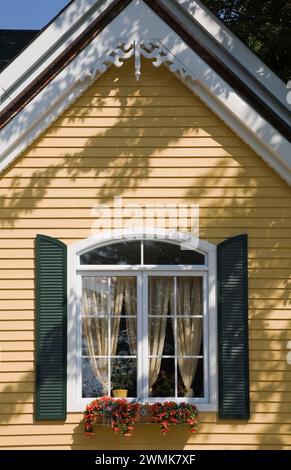 Bogenfenster mit dekorativen grünen Sturmläden auf gelber Schranke mit weißen Zierleisten im viktorianischen Stil im Sommer. Stockfoto