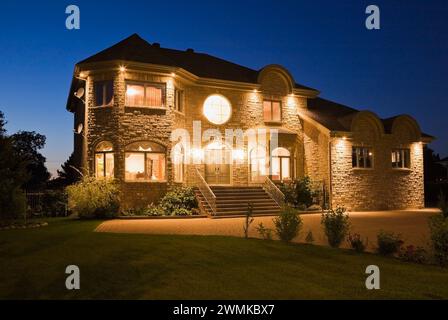 Luxuriöses zweistöckiges Haus aus braunem Stein mit landschaftlich gestaltetem Vorhof und gepflasterter Steineinfahrt beleuchtet in der Abenddämmerung. Stockfoto