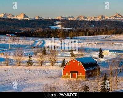 Rote Scheune in einem schneebedeckten Feld, das im warmen Licht des Sonnenaufgangs leuchtet, mit Ausläufern und schneebedeckten Bergen im Hintergrund und blauem Himmel, W... Stockfoto