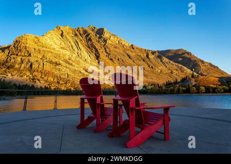 Glühender, farbenfroher Berg bei Sonnenaufgang mit zwei roten Stühlen auf einem Dock und blauem Himmel im Waterton Lakes National Park; Waterton, Alberta, Kanada Stockfoto