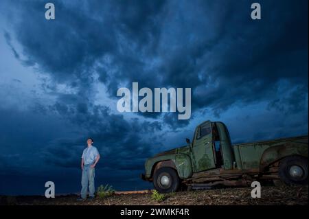 Junger Mann schaut auf die sich sammelnden Sturmwolken am Himmel in der Nähe eines 1951 International Harvester Pickups auf einem Bauernhof Stockfoto