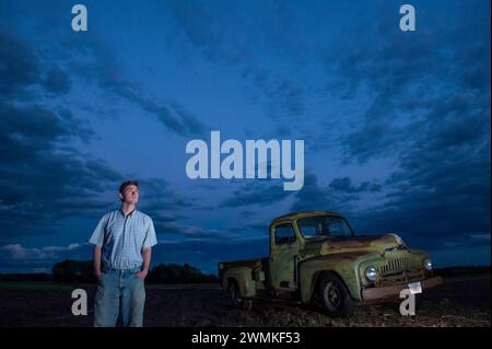 Junger Mann schaut auf die sich sammelnden Sturmwolken am Himmel in der Nähe eines 1951 International Harvester Pickups auf einem Bauernhof Stockfoto