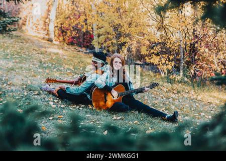 Gerahmte Ansicht eines verheirateten Ehepaares mit gemischter Rasse, das auf dem Gras sitzt und Gitarren spielt, lächelt und während einer Herbstfamilie für die Kamera posiert... Stockfoto