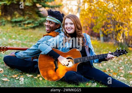 Nahaufnahme eines verheirateten Paares mit gemischter Rasse, das auf dem Gras sitzt und Gitarren spielt, lächelt und während eines Herbstes für die Kamera posiert... Stockfoto