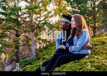 Porträt eines Paares gemischter Rassen, das auf einem grasbewachsenen Hügel sitzt und die Aussicht genießt, während eines Familienausflugs im Herbst in einer Stadt eine gute Zeit miteinander verbringt... Stockfoto