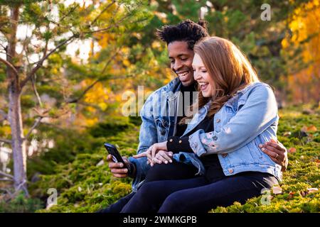 Nahaufnahme einer gemischten Rasse verheiratetes Paar, das lächelt und zusammen auf das Handy schaut, in der Dämmerung auf dem Gras sitzt, schöne Zeit miteinander verbringt... Stockfoto