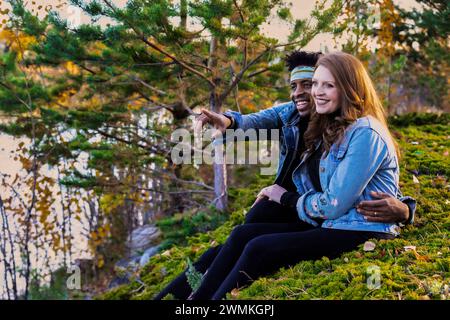 Porträt eines Paares gemischter Rassen, das auf einem grasbewachsenen Hügel sitzt und die Aussicht genießt, während eines Familienausflugs im Herbst in einer Stadt eine gute Zeit miteinander verbringt... Stockfoto