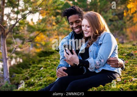 Nahaufnahme einer gemischten Rasse verheiratetes Paar, das lächelt und zusammen auf das Handy schaut, in der Dämmerung auf dem Gras sitzt, schöne Zeit miteinander verbringt... Stockfoto