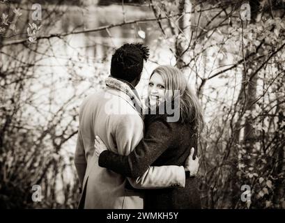 Blick von hinten auf ein Paar gemischter Rassen, das am Ufer eines Sees steht und sich umarmt, während die Frau zurückblickt und in die Kamera lächelt, ... Stockfoto