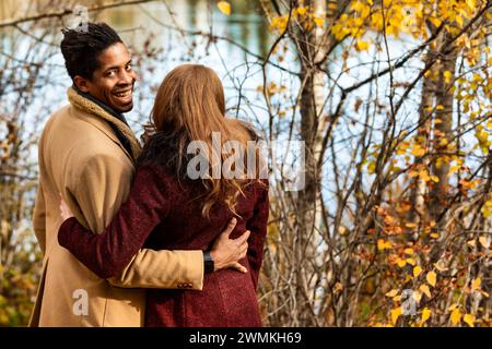 Blick von hinten auf ein gemischtes Paar, das am Ufer eines Sees steht und sich umarmt, während der Mann zurückblickt und den Camer anlächelt... Stockfoto