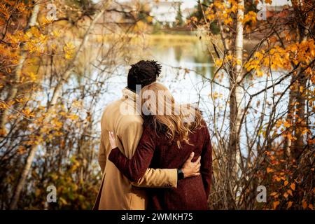 Blick von hinten auf ein Paar gemischter Rassen, das am Ufer eines Sees steht und sich umarmt, während es im Herbst gute Zeit miteinander verbringt... Stockfoto