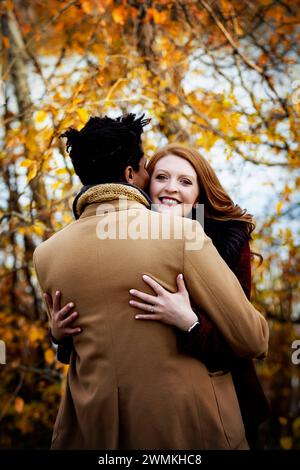 Blick von hinten auf ein Paar gemischter Rassen, das sich umarmt, während die Frau in die Kamera lächelt und während eines herbstfami Zeit miteinander verbringt... Stockfoto