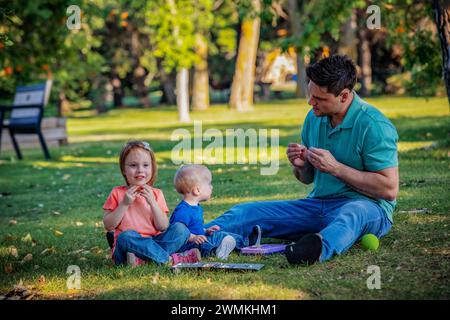 Der Vater verbringt Zeit und die Gebärdensprache mit seinem kleinen Sohn, der an dem Down-Syndrom leidet, während seine Tochter mitspielt, in einem Stadtpark, der... Stockfoto