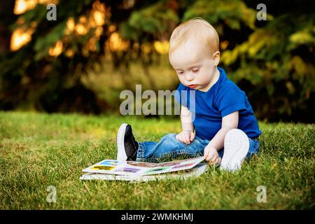 Junge mit Down-Syndrom, der an einem warmen Herbstnachmittag in einem Stadtpark sitzt und sich ein Bilderbuch über Gras anschaut; Leduc, Alberta, Kanada Stockfoto