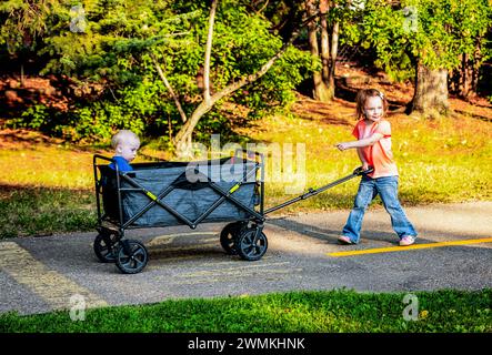 Vorschulmädchen zieht ihren jüngeren Bruder mit Down-Syndrom in einem Wagen auf einem Weg in einem Stadtpark während eines warmen Herbstabends Stockfoto
