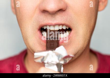 Mund eines Menschen, der Schokolade vor blauem Hintergrund isst. Der Mund eines Mannes in Nahaufnahme mit weißen Zähnen, die dunkle Schokolade beißen. Stockfoto