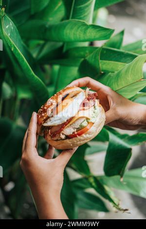 Die Hände halten Burger-Bagels, gefüllt mit Gemüse, Hühnchen, Eiern, dahinter stehen Pflanzen Stockfoto