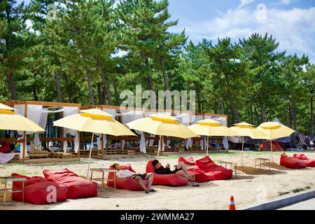 Yangyang County, Südkorea - 30. Juli 2019: Strandbesucher entspannen sich auf roten Sitzsackstühlen unter gelben Sonnenschirmen am Sandstrand von Hyangho Beach, W Stockfoto