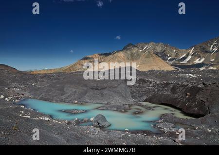 Der türkisfarbene Gletschersee liegt inmitten dunkler Moränen unter einer schroffen Bergkette und blauem Himmel Stockfoto