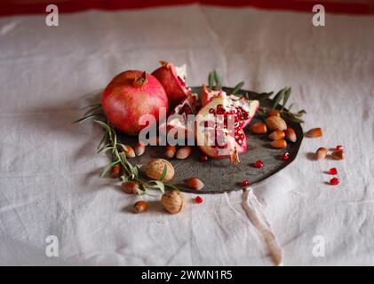 Bild des Stilllebens mit türkischem Granatapfel und Olivenzweig auf alter Retro-Platte. Dunkler Holzhintergrund, antike Kupferplatte. Frische reife ganze Granatäpfel, geöffnete Granatäpfel und Samen Stockfoto