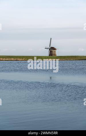 Einsame traditionelle Strohdachmühle am Dutch Lake in der Abenddämmerung. Halb versteckt hinter einem grünen Deich mit Blick auf das ruhige Wasser einer überfluteten Wiese Stockfoto